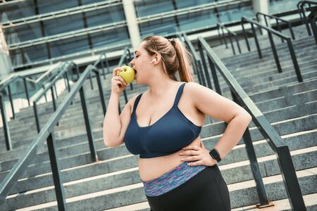 Healthy food. Plus size woman in sport clothing eating green apple while standing on stairs outside.の写真素材