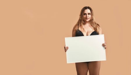 Your text here. Pretty and confident plus size young woman in black lingerie holding empty blank board while posing in studio on brown background.の写真素材