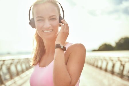 Feeling happy. Beautiful young woman in headphones listening music and looking at camera with smile while standing on the bridgeの写真素材