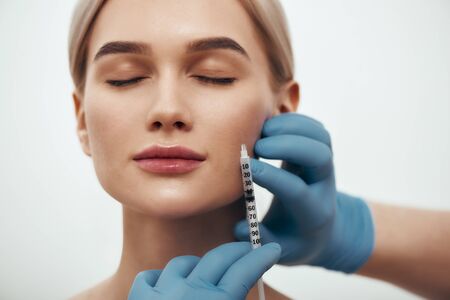 Wanna be perfect. Portrait of young pretty woman keeping eyes closed while doctors hand in blue medical gloves making an injection in her faceの写真素材