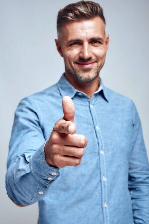 Smiling handsome man in blue shirt pointing his finger and smiling at camera while standing against...