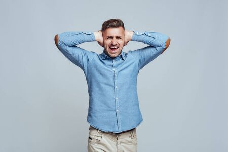 Depressed man closing ears with both hands and screaming while standing against greyの写真素材