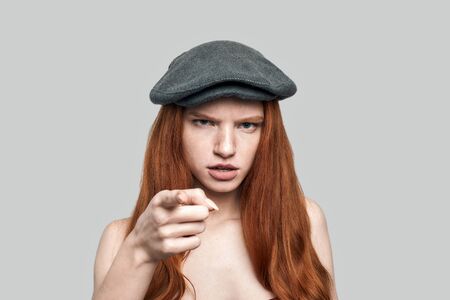 Portrait of young rude redhead woman in headgear looking at camera and gesturing while standing against greyの写真素材
