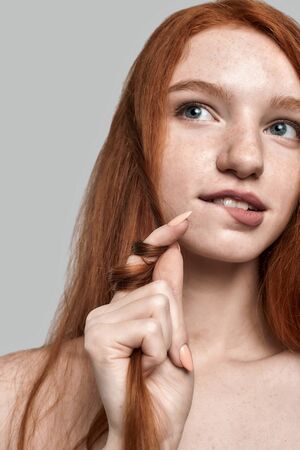 Portrait of young and cute redhead woman playing with her hair and thinking about something while standing against greyの写真素材