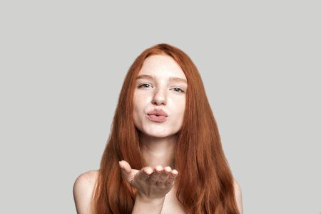 Sending you my love. Portrait of cute young lady with red hair blowing a kiss at the camera while standing against grey backgroundの写真素材