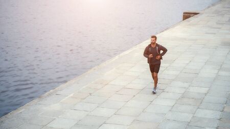 I am starting new day with sport. Young and handsome bearded man in sports clothing running along the riverbankの写真素材