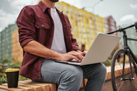Working outdoors. Cropped photo of a man with stubble in casual clothes working on laptop and drinking coffee while sitting on the bench near his bicycleの写真素材
