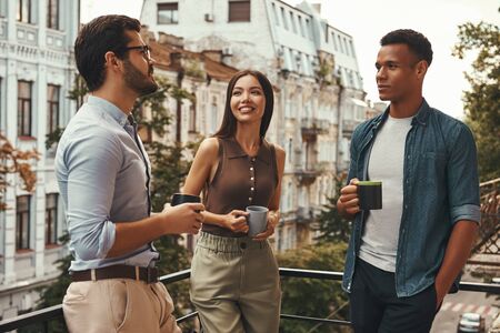 Coffee break. Young cheerful colleagues in casual wear holding cups and talking with each other...