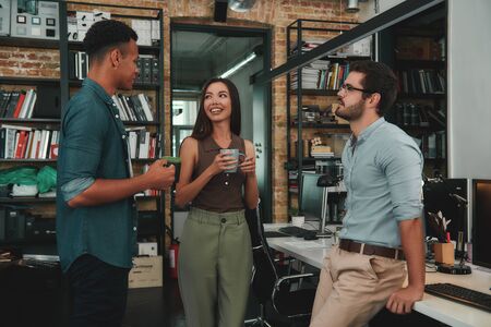 Nice coffee! Young smiling colleagues in casual wear holding cups and talking about something while standing in modern officeの写真素材