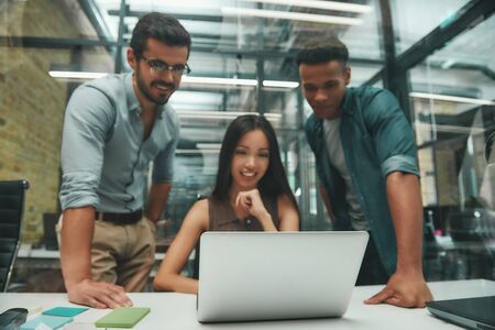 Using modern technologies. Group of three young and cheerful employees looking at screen of laptop...