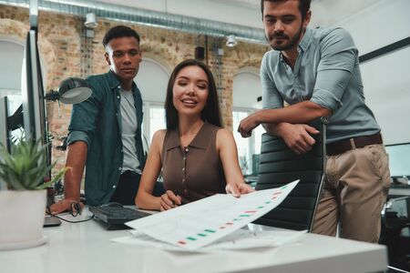 Coworking space. Group of young and cheerful three people in casual clothes analyzing data and...