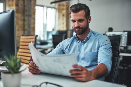 Great work Concentrated bearded engineer in formal wear looking at blueprint and smiling while sitting in the modern officeの写真素材