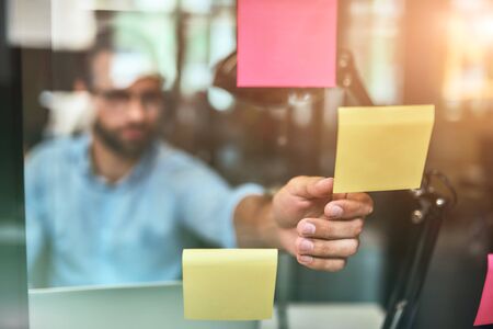 Business ideas. Blurred photo of bearded man in eyeglasses using colorful stickers while sitting in front of glass wallの写真素材