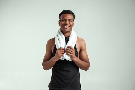 It was great workout. Handsome african man with towel on shoulders looking at camera and smiling while standing against grey backgroundの写真素材