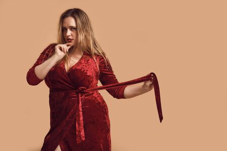 Feeling beautiful. Plus size young woman in velour dress touching her chin and looking at camera while standing in studioの写真素材