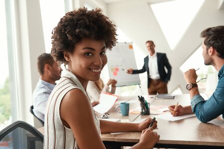 Young afro american woman is looking at camera and smiling while her boss standing near whiteboard...