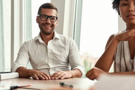 Happy working day. Young and handsome man in eyeglasses holding cup of coffee and looking away with smile while sitting with colleagues at the office tableの写真素材