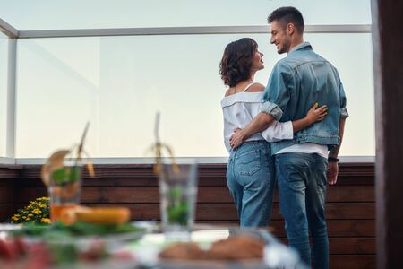Romantic evening. Back view of young couple in casual clothes are looking at each other while standing on rooftop terraceの写真素材