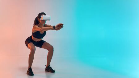Full length of young sporty woman in sports clothing doing squat while wearing virtual reality glassesの写真素材