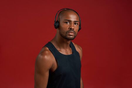 Portrait of handsome sporty afro american man in headphones looking at camera while standing against red backgroundの写真素材