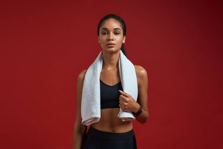 After workout. Beautiful afro american woman with towel on her shoulders looking at camera while standing against red backgroundの写真素材