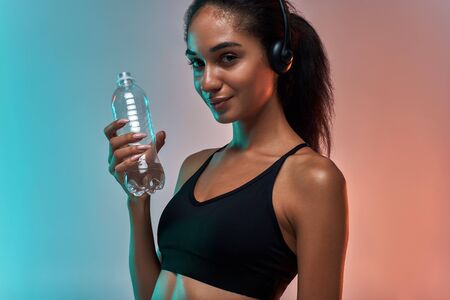 Enjoying music. Portrait of cute and sporty woman in headphones holding bottle of water and looking at camera with smile while standing against colorful backgroundの写真素材