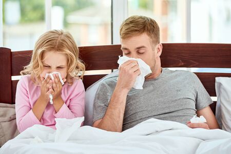 Sick family. Father and daughter suffering from flu or cold and wiping noses while lying in bed together at home. Virus diseaseの写真素材