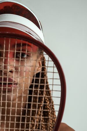 Winner. Close up portrait of beautiful young african woman holding tennis racket in front of her face and looking at camera while standing against grey backgroundの写真素材