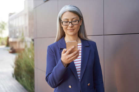Portrait of a confident mature business woman in classic wear using smartphone while standing...