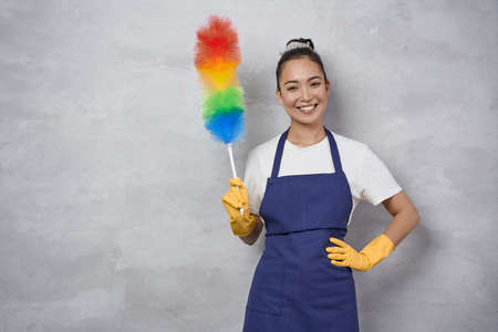 Happy young woman, cleaning lady in uniform holding colorful microfiber duster and smiling at camera...