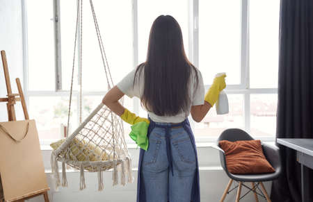 Housekeeping and cleaning service concept. Back view of a young woman, cleaning lady in yellow rubber gloves holding rag and spray while cleaning big panoramic window at homeの写真素材