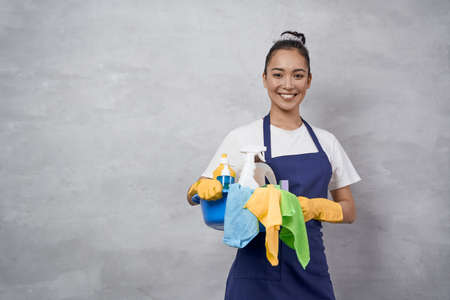 Young happy woman, female cleaner in uniform and rubber gloves holding bucket of cleaning supplies and smiling at camera, standing against grey wallの写真素材
