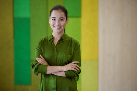 Portrait of a young beautiful female office worker keeping arms crossed, looking at camera and smiling while standing in the modern coworking spaceの写真素材