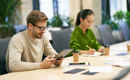 Working together. Young smiling caucasian man wearing eyeglasses using digital tablet while sitting at desk with his female colleague in the modern coworking spaceの写真素材