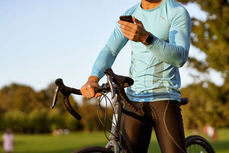 Professional cyclist in sportswear using his smartphone and resting after training while standing with his bike in the park on a sunny dayの写真素材