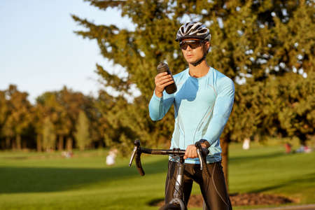 Taking a break. Young athletic man in sportswear and protective helmet standing with his bike in park, drinking water and resting after bicycle rideの写真素材
