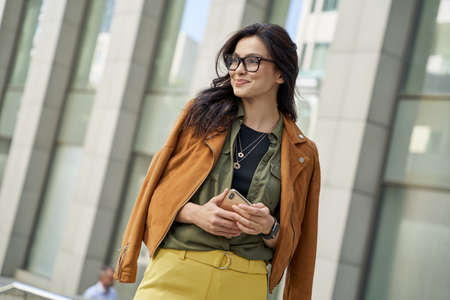 Young stylish woman holding smartphone, looking aside and smiling while standing against blurred...