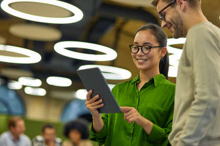 Young cheerful asian woman showing something on digital tablet to her male colleague while standing...