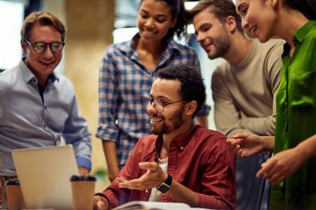 Group of happy multiracial business people looking at laptop screen and discussing work or project results while standing in the modern office or coworking spaceの写真素材