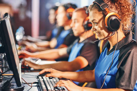 Side view of a happy mixed race girl, female cybersport gamer wearing headphone sitting in one row with team, participating in esports tournamentの写真素材