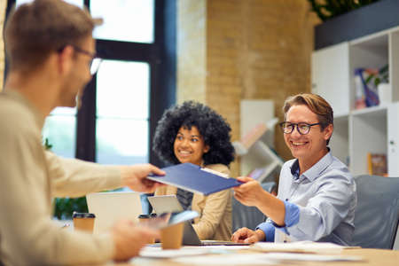 Cheerful man giving a folder with documents to his colleague and smiling while working together in...