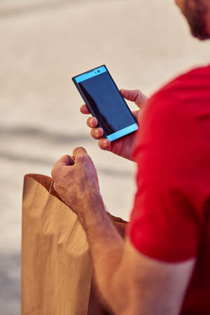 Rear view of a male courier using mobile app while delivering paper eco bag with food from shop or...