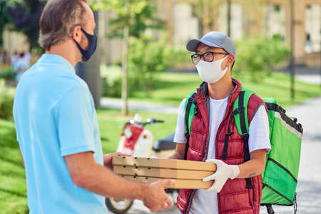 Young asian courier wearing face protective mask and gloves giving boxes with pizza to customer while standing on a sunny street. Food delivery during coronavirus pandemicの写真素材