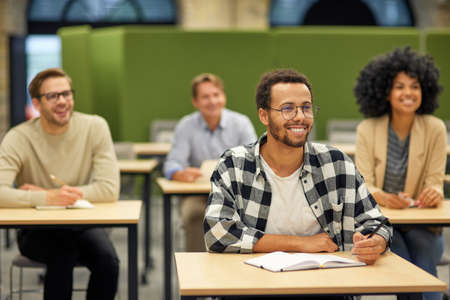 Young cheerful mixed race male office worker sitting listening to coach during seminar or training...