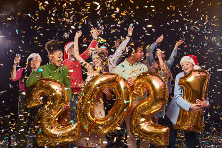 Group of young happy multiracial people celebrating New Years Eve, holding golden foil balloons in...