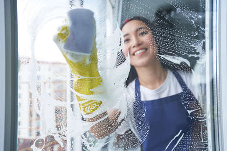 Best cleaning service. Cheerful young woman smiling while cleaning the window, glass surface using spongeの写真素材