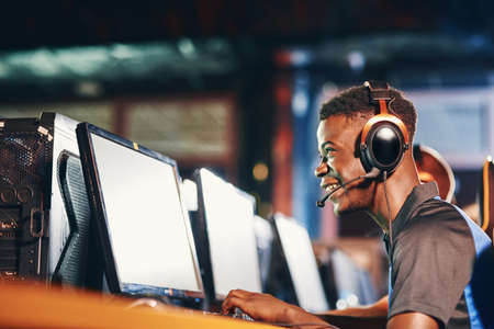 Side view of a happy excited african guy, professional gamer wearing headphones participating in eSport tournament, looking at PC screen while sitting in gaming clubの写真素材