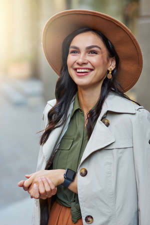 Vertical shot of attractive happy stylish woman wearing grey coat and hat looking aside and smiling while standing on the city street, enjoying walking on warm autumn dayの写真素材