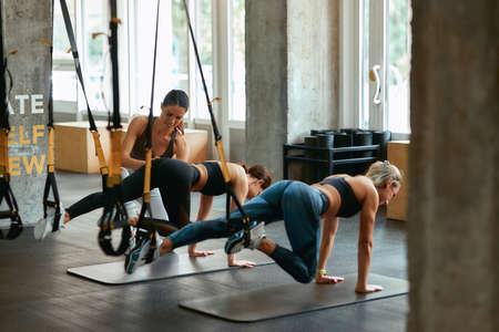 Developing physical strength. Two young athletic girls exercising with trx fitness straps with female personal trainer at gymの写真素材