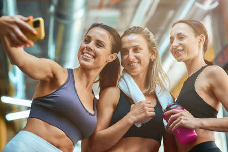 Happy moments. Group of three young beautiful and cheerful sportive women taking selfie on smartphone while resting after workout, exercising together at gymの写真素材